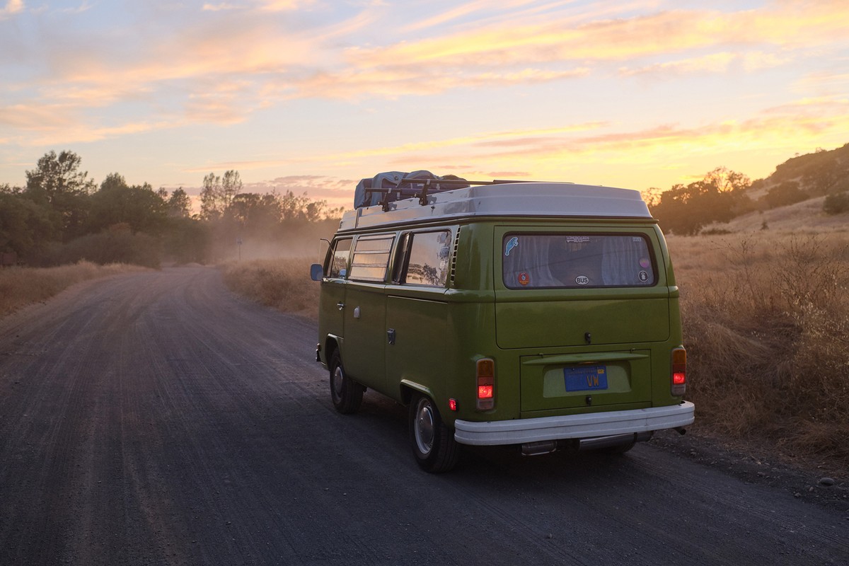 Rear view of a vintage green Volkswagen camper van driving on a dirt road at sunset, with luggage on top, in a rural setting.