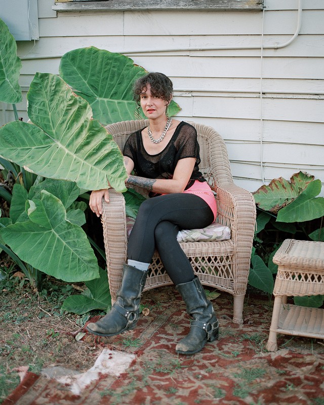 Woman with a tattoo sits in a wicker chair wearing black boots and clothing, in front of a house with large leafy plants.