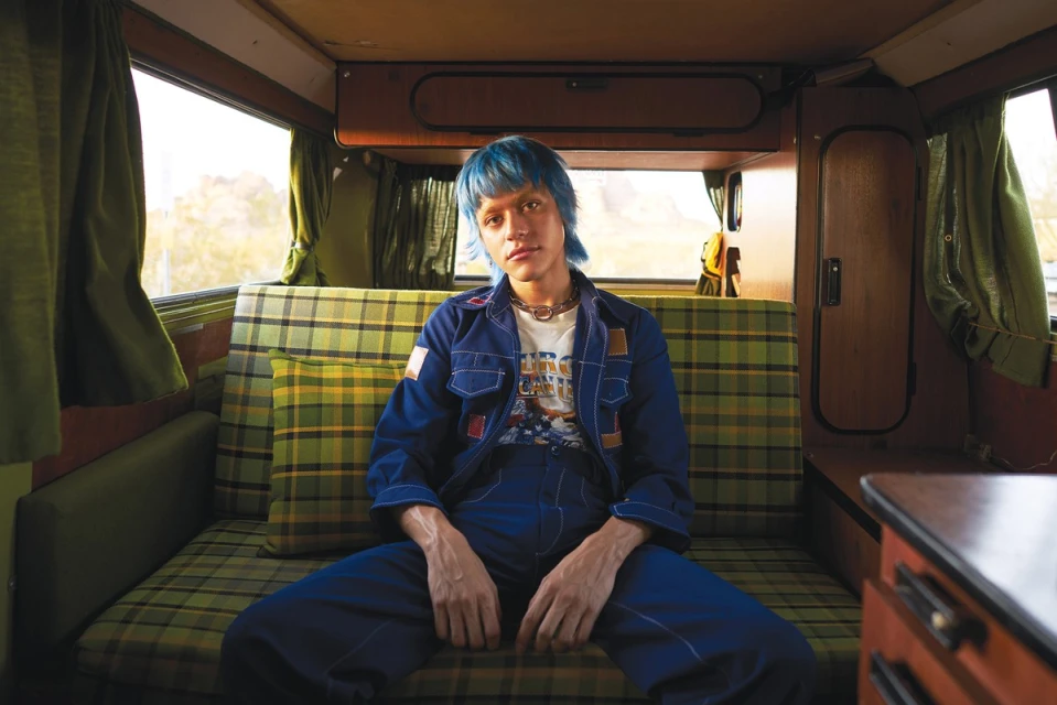 Adult with blue hair sits in a vintage camper van on a plaid patterned seat wearing a blue denim jumpsuit and white graphic t-shirt.