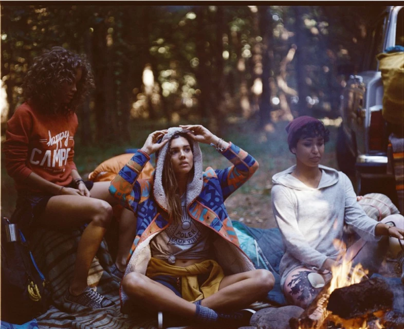 Three women in outdoor fashion, camping attire, sitting near a campfire in a wooded setting, wearing casual tops and outer layers.