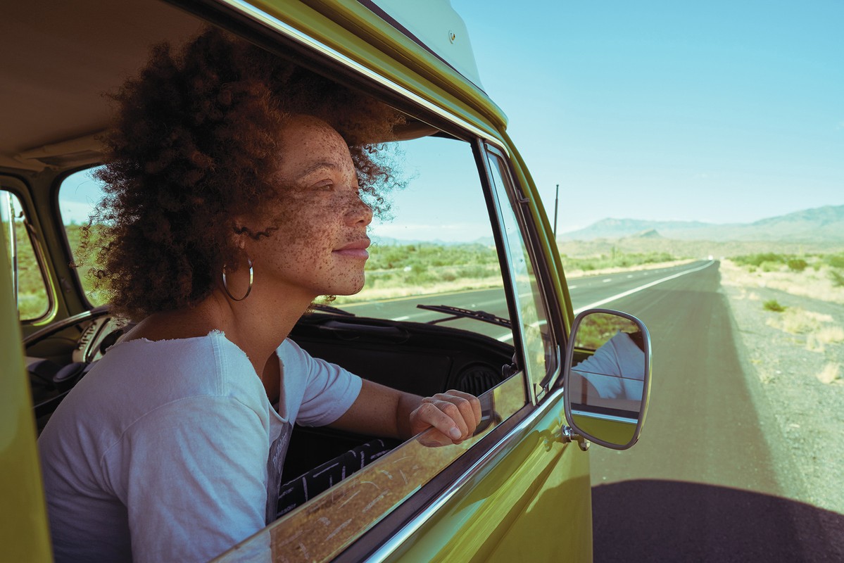 A curly-haired woman with freckles leans out the window of a retro olive-green van on an open road with mountains in the background.