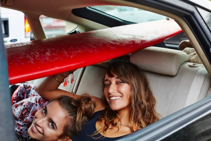 Two smiling young women lying in the back seat of a car beneath a red surfboard