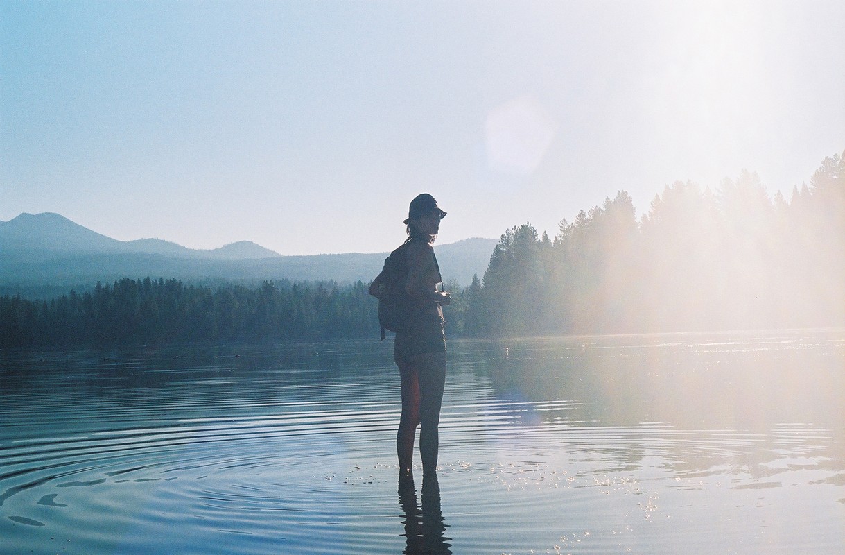 Woman stands in a lake with a forest and mountains in the background, clear skies, wearing a hat and carrying a backpack.
