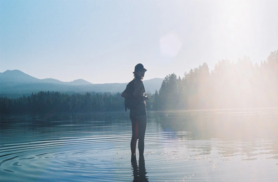 Woman stands in a lake with a forest and mountains in the background, clear skies, wearing a hat and carrying a backpack.