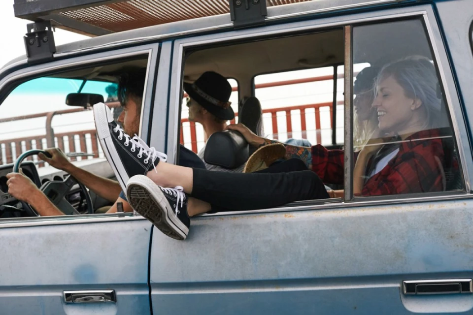 Group of four people traveling in a blue SUV with legs out the window, showing Converse shoes, driving on a bright, sunny day.