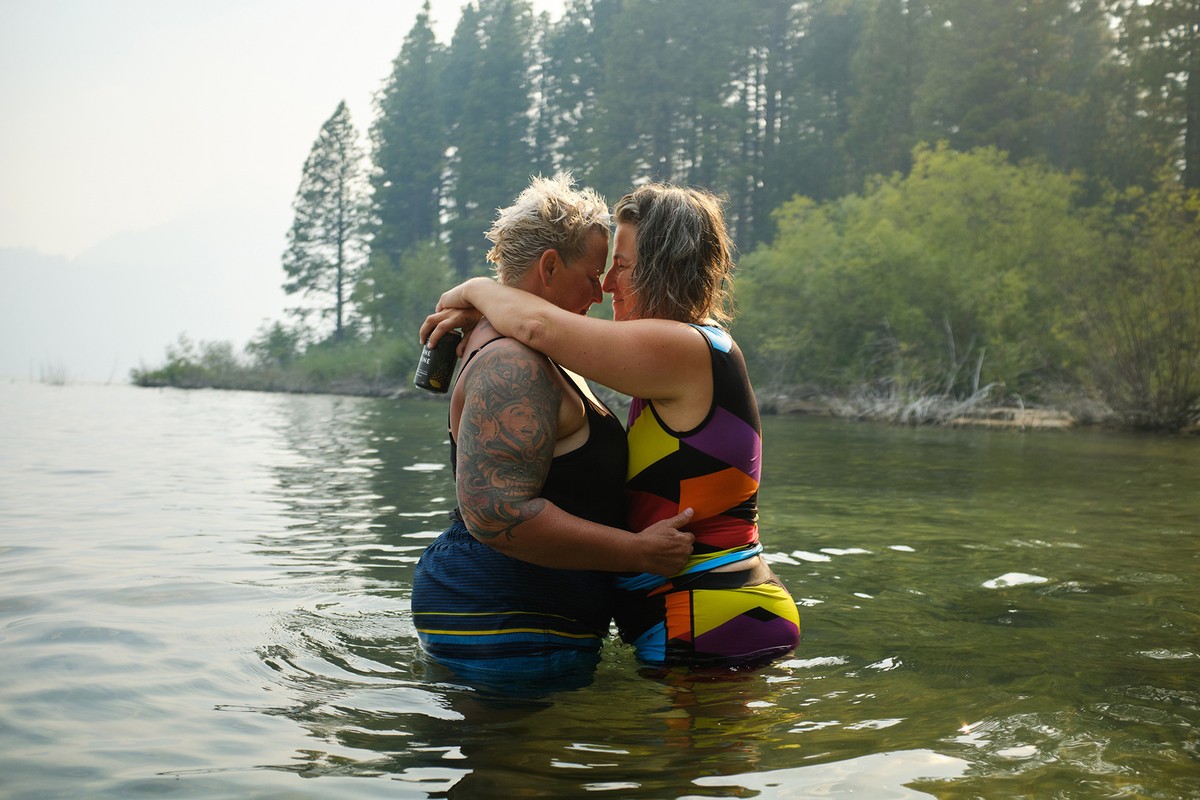 Two women embrace in lake water surrounded by a forest of green trees in an eye-level medium shot capturing an intimate, outdoor moment.