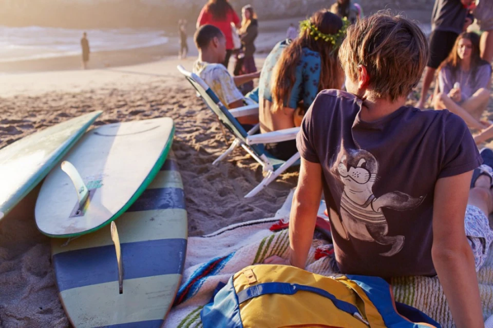 People relax on a beach with surfboards during the day. The warm sunlight illuminates the scene, creating a relaxed atmosphere.