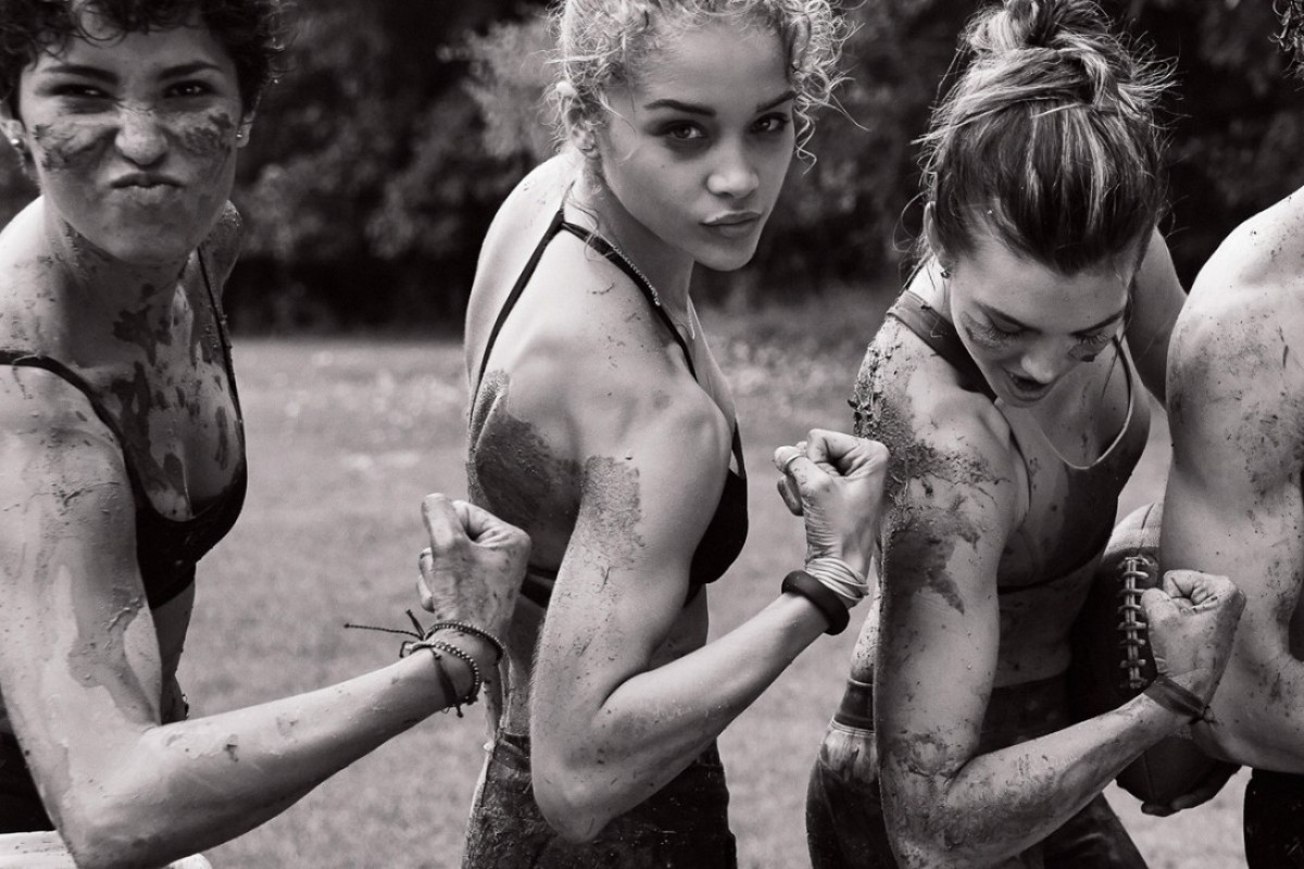 Black and white shot of four women flexing their biceps, covered in mud, outdoors in a field with trees in the background.