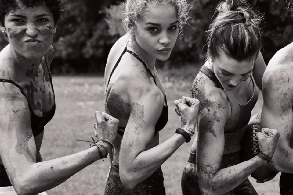 Black and white shot of four women flexing their biceps, covered in mud, outdoors in a field with trees in the background.