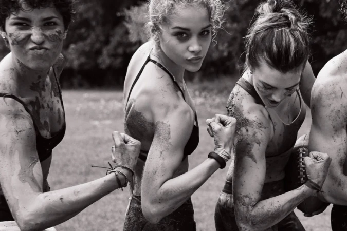 Black and white shot of four women flexing their biceps, covered in mud, outdoors in a field with trees in the background.