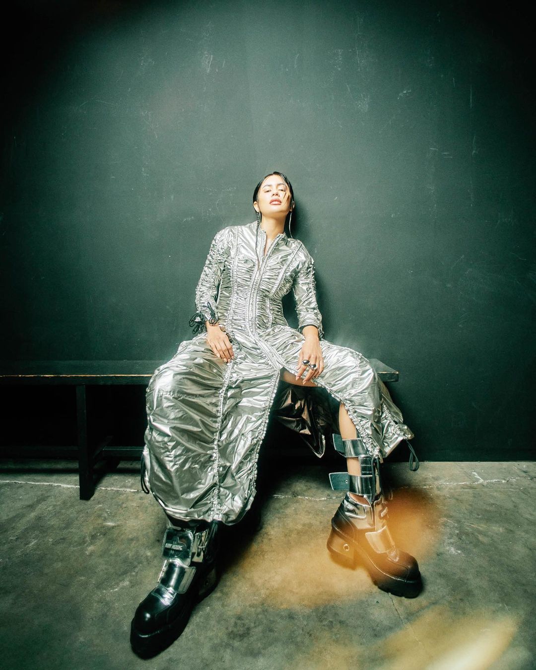 Woman in futuristic silver dress seated on bench. Fashion-forward attire, glossy texture, dark backdrop, and avant-garde footwear.