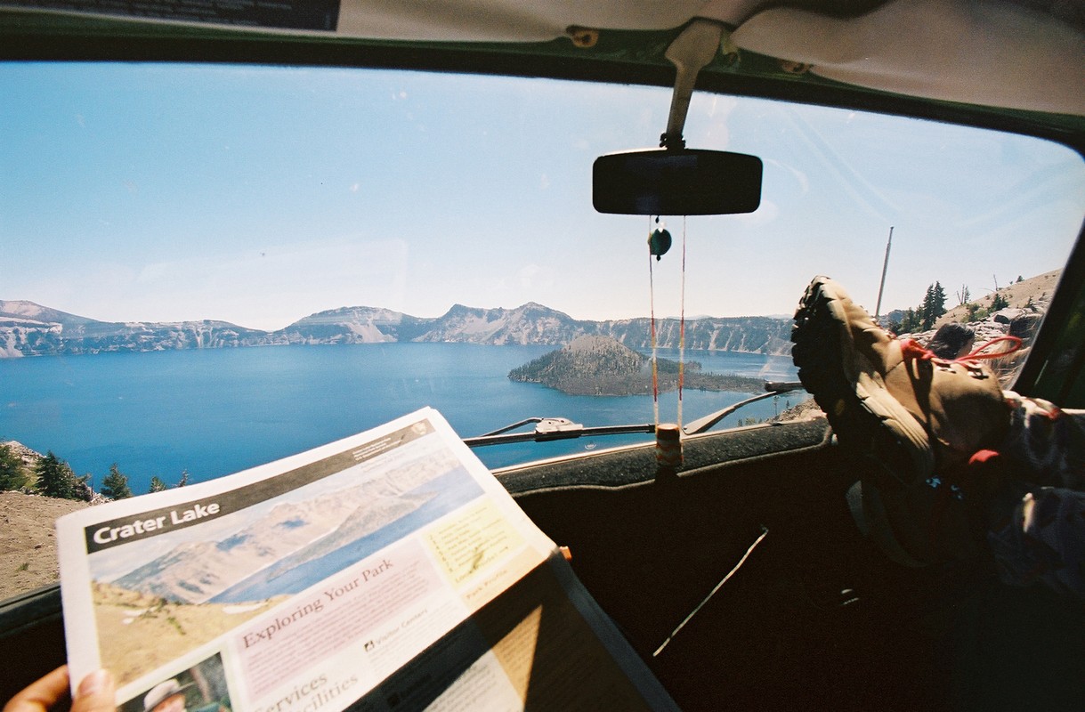 View from inside a car of Crater Lake, Oregon, with a travel brochure and a hiking boot visible in the foreground.