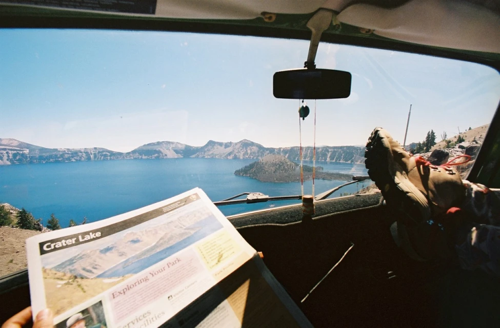 View from inside a car of Crater Lake, Oregon, with a travel brochure and a hiking boot visible in the foreground.