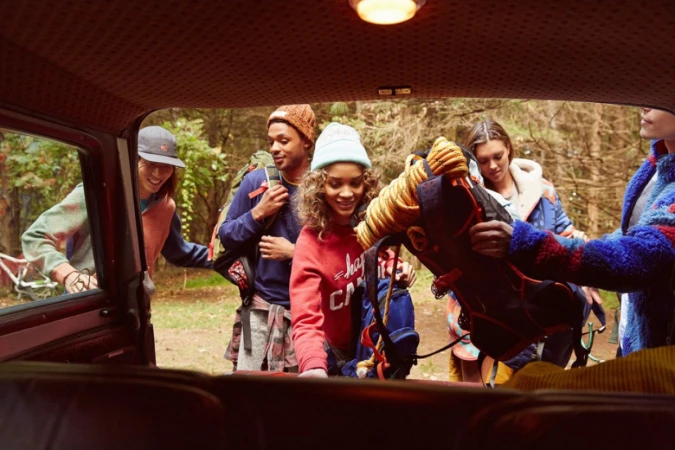 A group of five people outside of a vehicle in a forest unpack a backpack, ready for a camping or hiking trip in the outdoors.