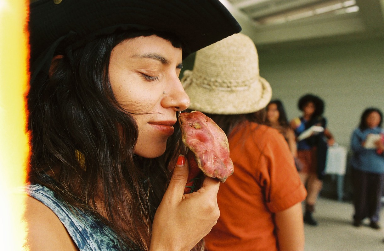Woman in hat with eyes closed, holding produce in outdoor setting with group. Natural light highlights her features and the scene.