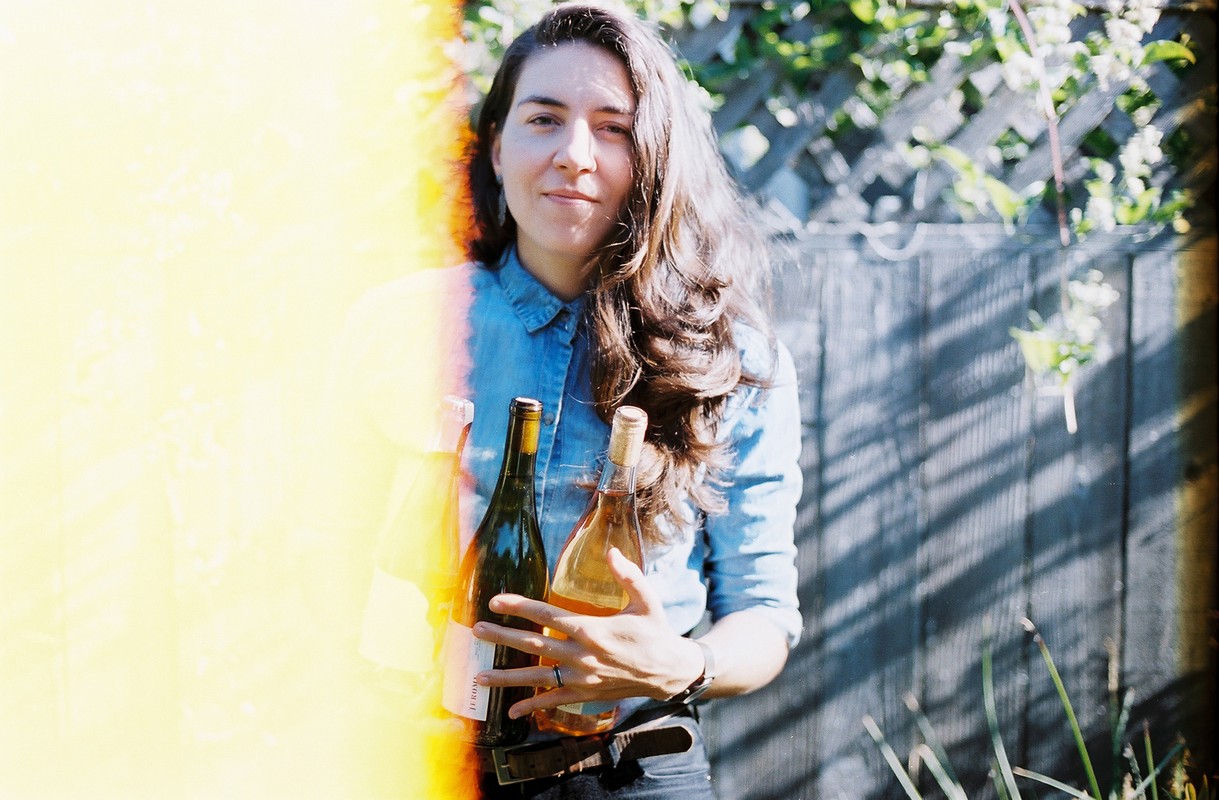 Young woman in denim shirt outdoors holding two bottles of wine in front of wooden fence and vine-covered trellis, with sunny side-flare.