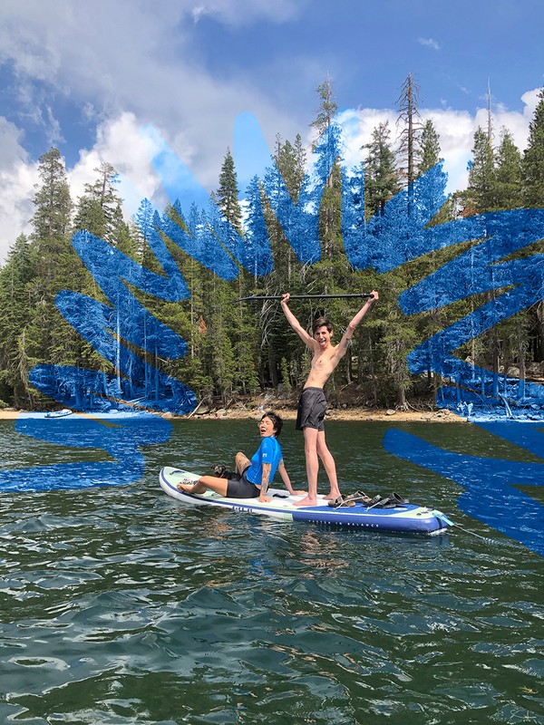 Two shirtless young men paddle boarding on a lake surrounded by trees, clear sky with clouds and water reflections.