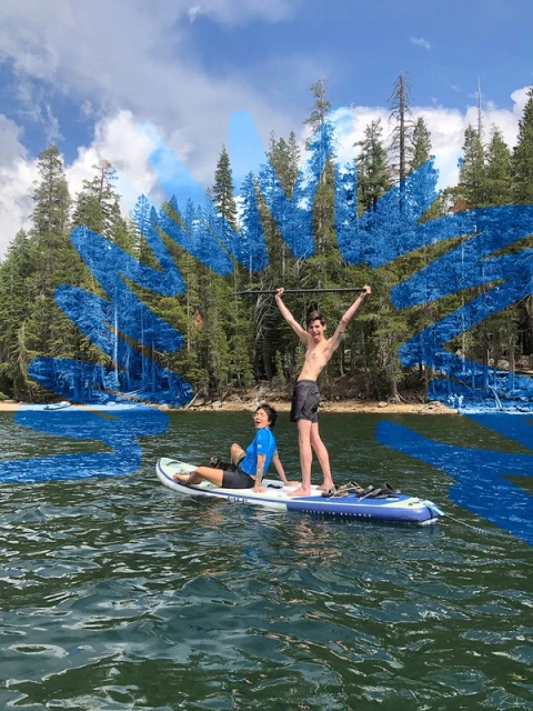 Two shirtless young men paddle boarding on a lake surrounded by trees, clear sky with clouds and water reflections.