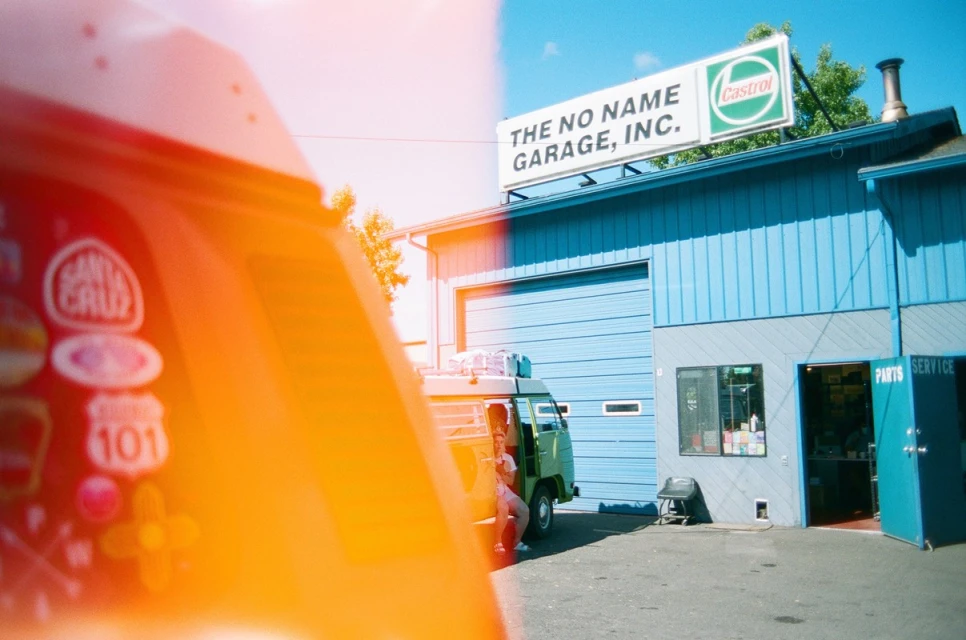 Exterior shot of a vintage Volkswagen van parked outside The No Name Garage, Inc., with a Castrol sign on the building.