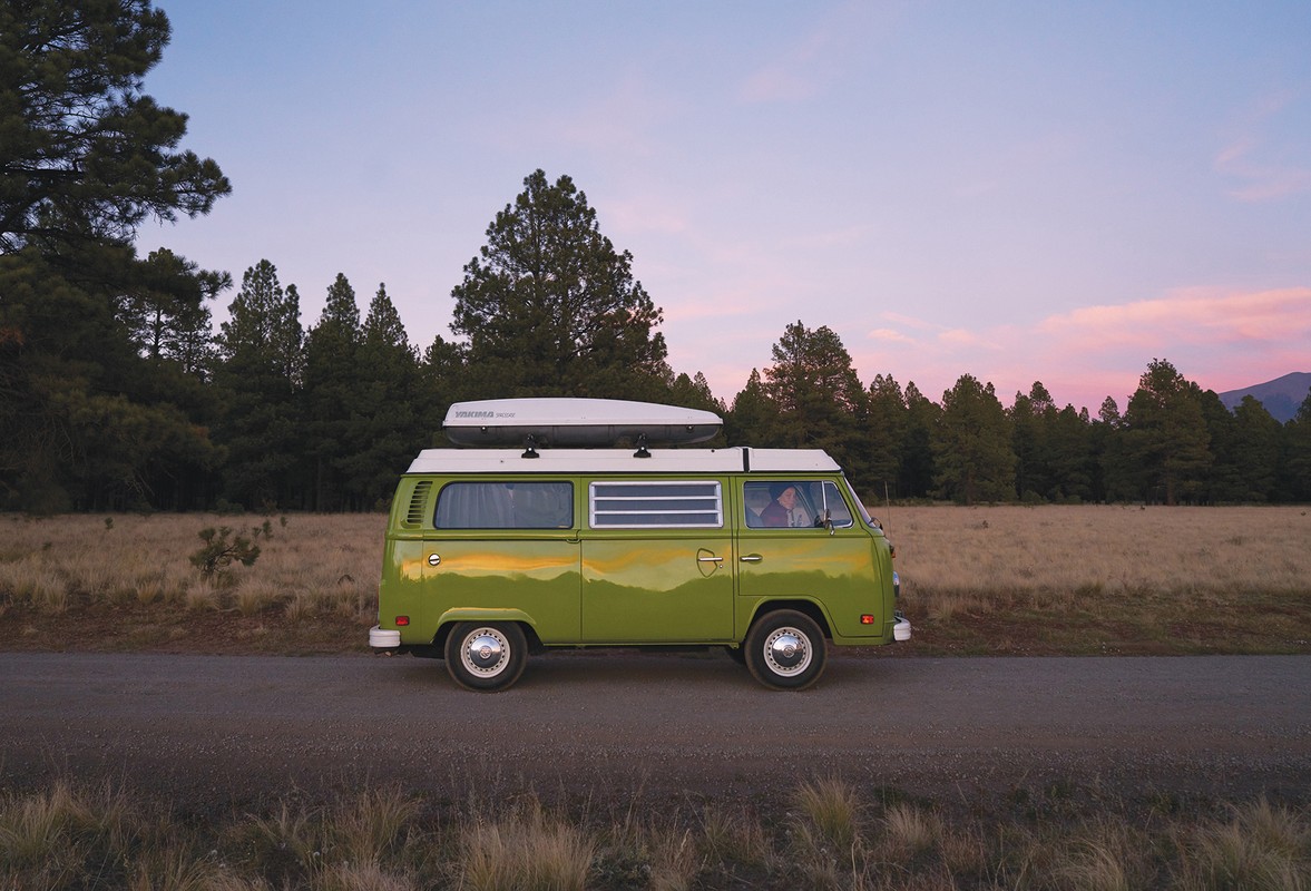 Green vintage Volkswagen bus parked in a grassy field with tall trees and a pink sunset sky, evoking a sense of adventure.