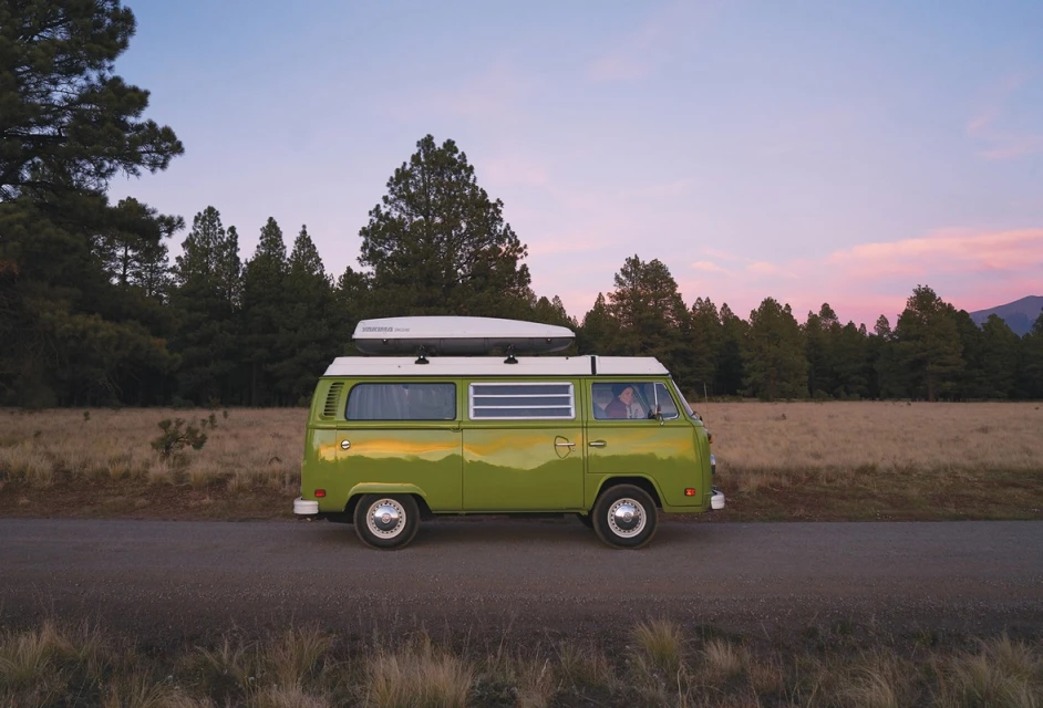 Green vintage Volkswagen bus parked in a grassy field with tall trees and a pink sunset sky, evoking a sense of adventure.