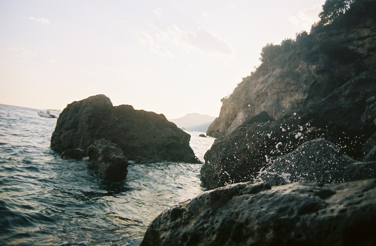 Scenic view of rocky shoreline with cliffs, water, and boulders at sunset. Splashing waves create a dynamic coastal landscape.