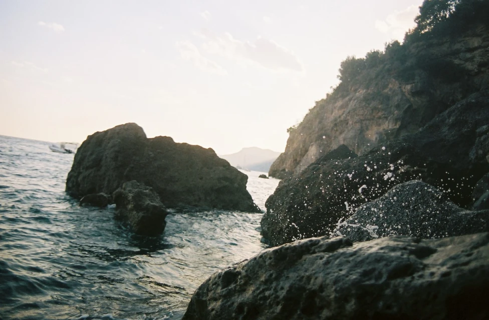 Scenic view of rocky shoreline with cliffs, water, and boulders at sunset. Splashing waves create a dynamic coastal landscape.
