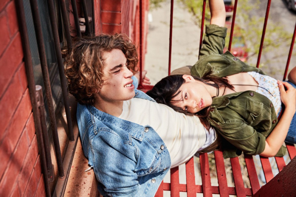 Two young adults lounge on a red-painted metal fire escape. The woman rests her head on the man's shoulder, wearing denim jackets.