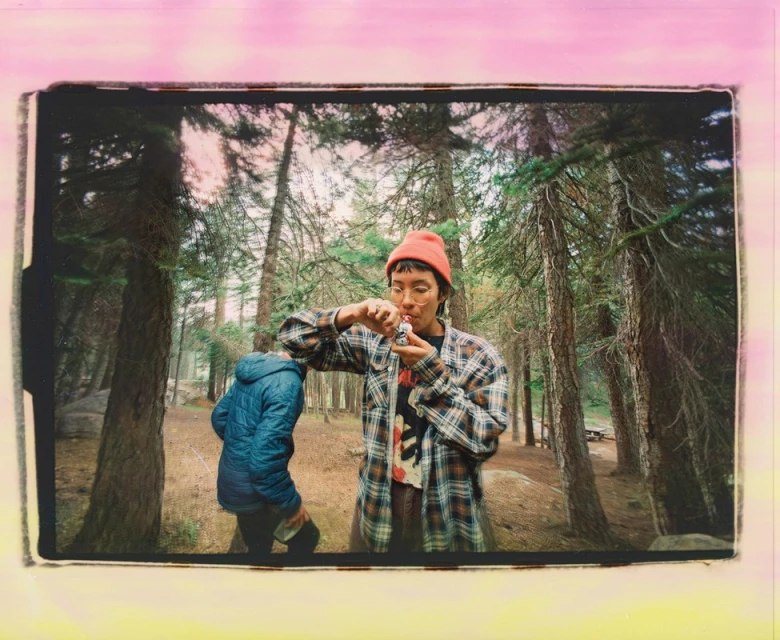 Two people outdoors in a forest with tall trees, one holding a small pipe, wearing a plaid shirt and orange beanie, sunny lighting.