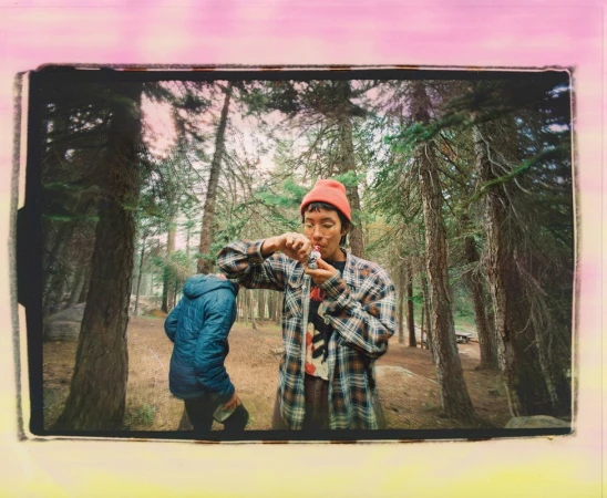 Two people outdoors in a forest with tall trees, one holding a small pipe, wearing a plaid shirt and orange beanie, sunny lighting.