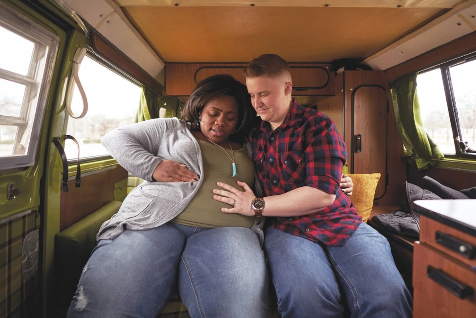 Two people inside a van. One person is touching the stomach of the other person. They are both sitting and smiling softly.