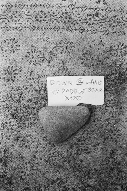 Close-up of a heart-shaped stone on patterned ground, accompanied by a handwritten note stating, 'Down @ Lake w/ Paddleboard XOXO'.