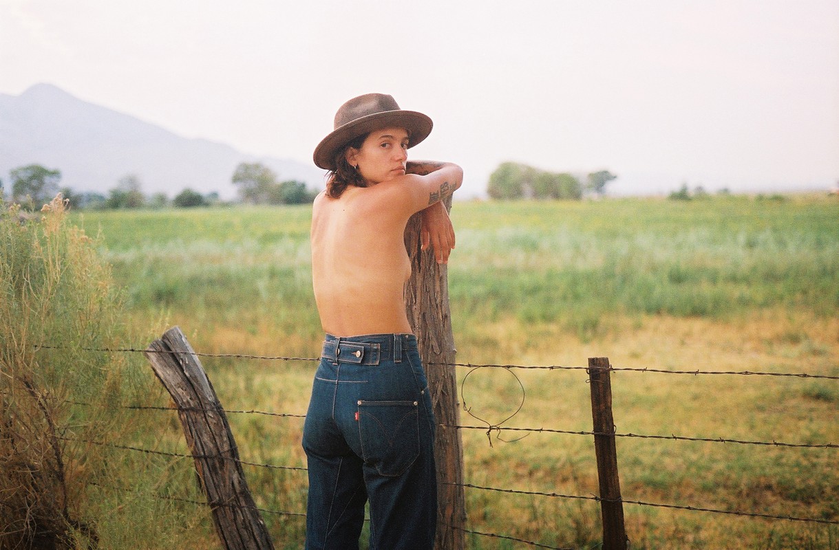 Woman in jeans and hat poses by a barbed wire fence in a rural setting with mountains in the background on a bright day.