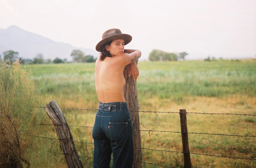 Woman in jeans and hat poses by a barbed wire fence in a rural setting with mountains in the background on a bright day.