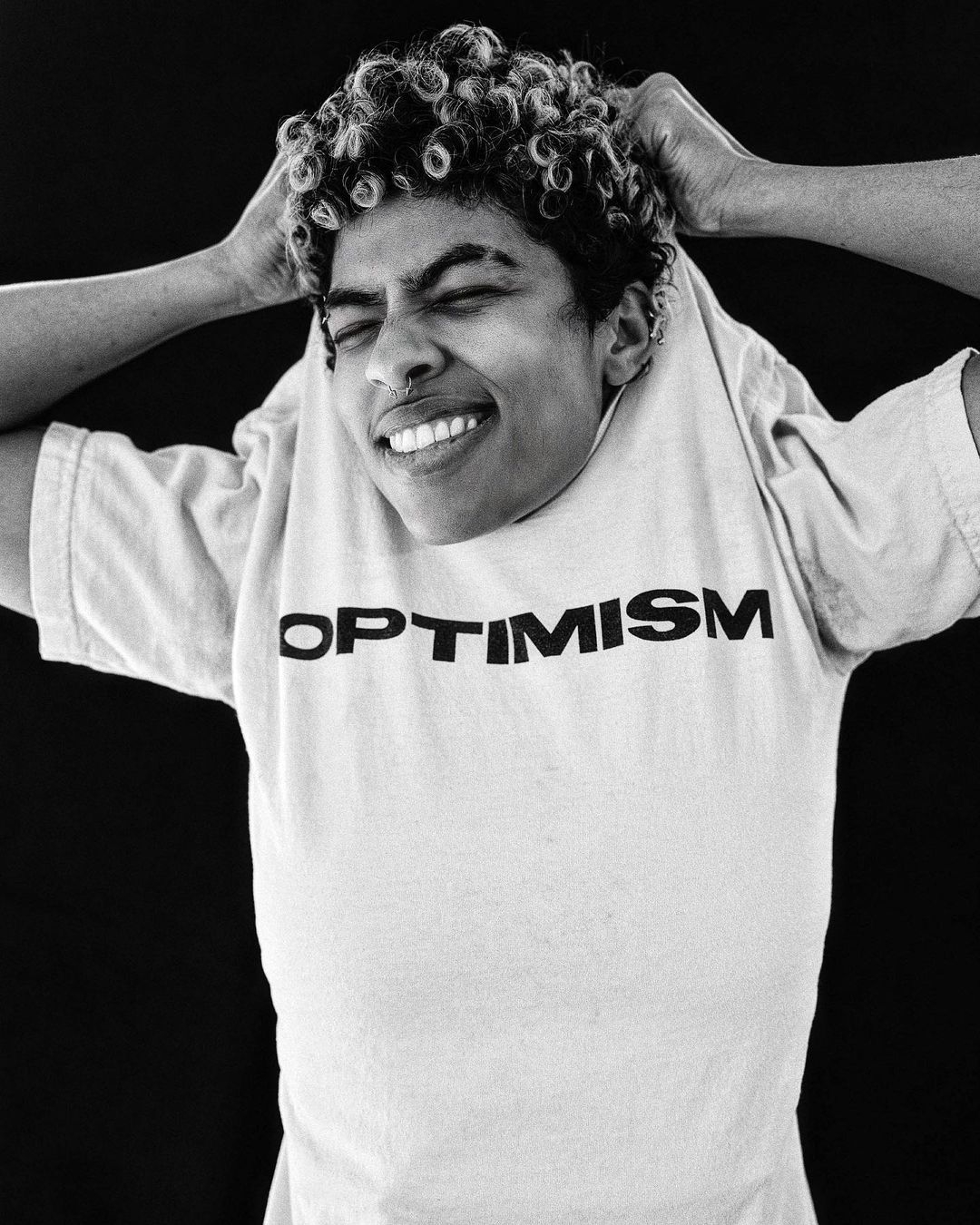 Portrait of a smiling young adult with curly hair, wearing a white 'Optimism' t-shirt against a plain black backdrop in a studio setting.