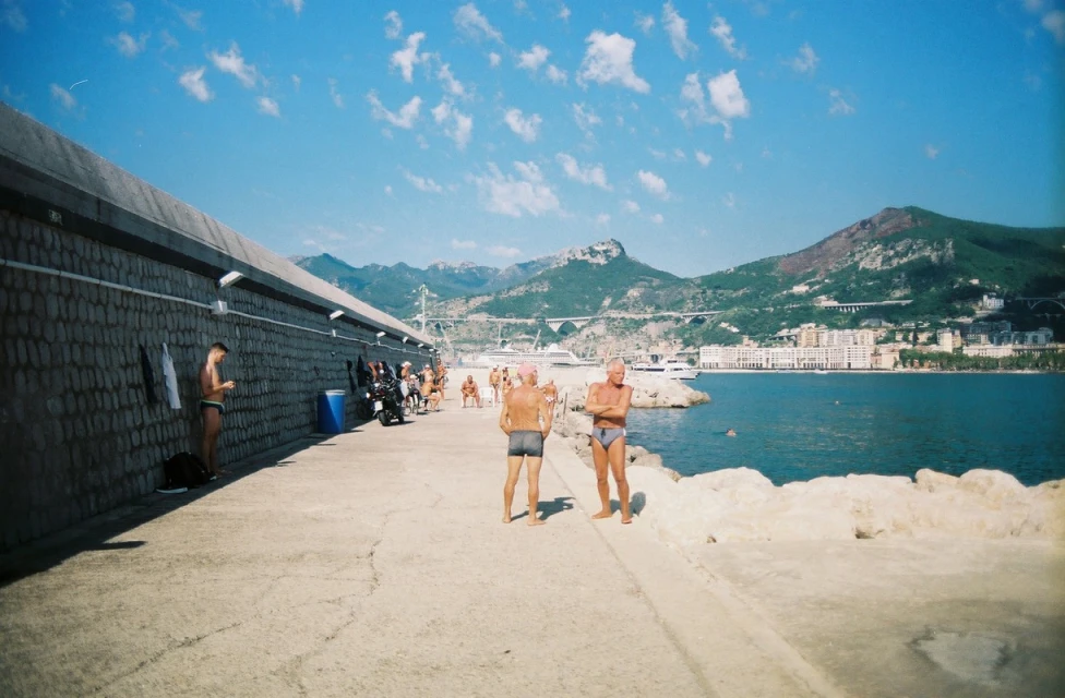 Two elderly men stand on a concrete pier by the sea with mountains and a city in the background, under a bright blue sky.
