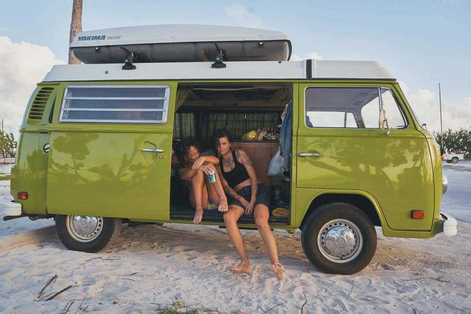 Two tattooed adult females in a retro green and white camper van parked on a beach, showcasing a lifestyle aesthetic with a vintage vibe.