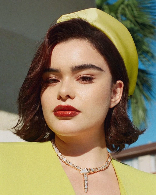 Close-up of a woman with short brown hair, red lipstick, and a yellow headband and jacket, wearing a white and gold necklace. 