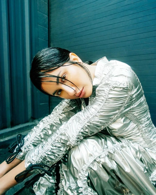 A woman in a metallic dress poses seated against a blue corrugated wall, looking at the camera with a neutral expression.