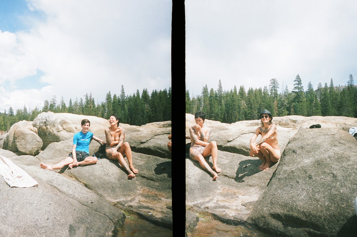 Two women pose on rocks beside a river with forest backdrop, bathed in natural light on a sunny day, capturing candid summer leisure.