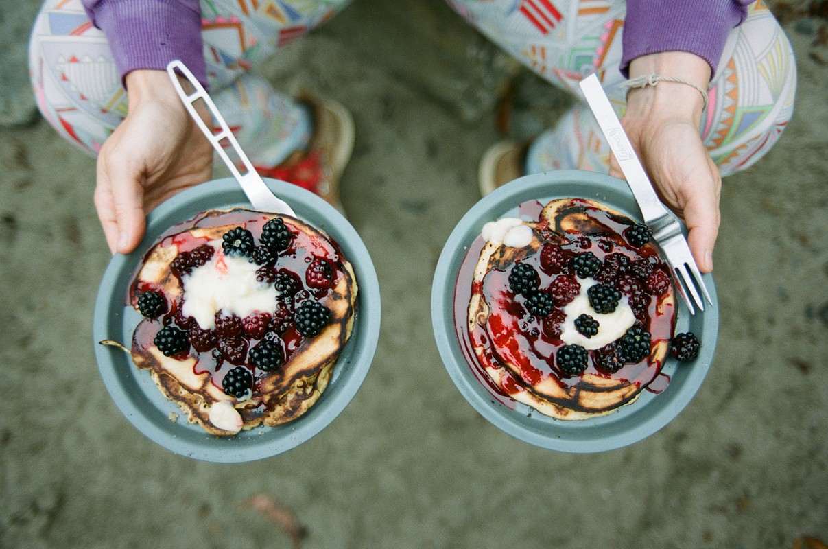 Overhead shot of two plates of pancakes topped with blackberries, cream, and syrup, held by a person wearing patterned leggings.