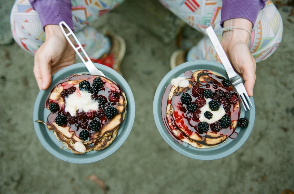 Overhead shot of two plates of pancakes topped with blackberries, cream, and syrup, held by a person wearing patterned leggings.