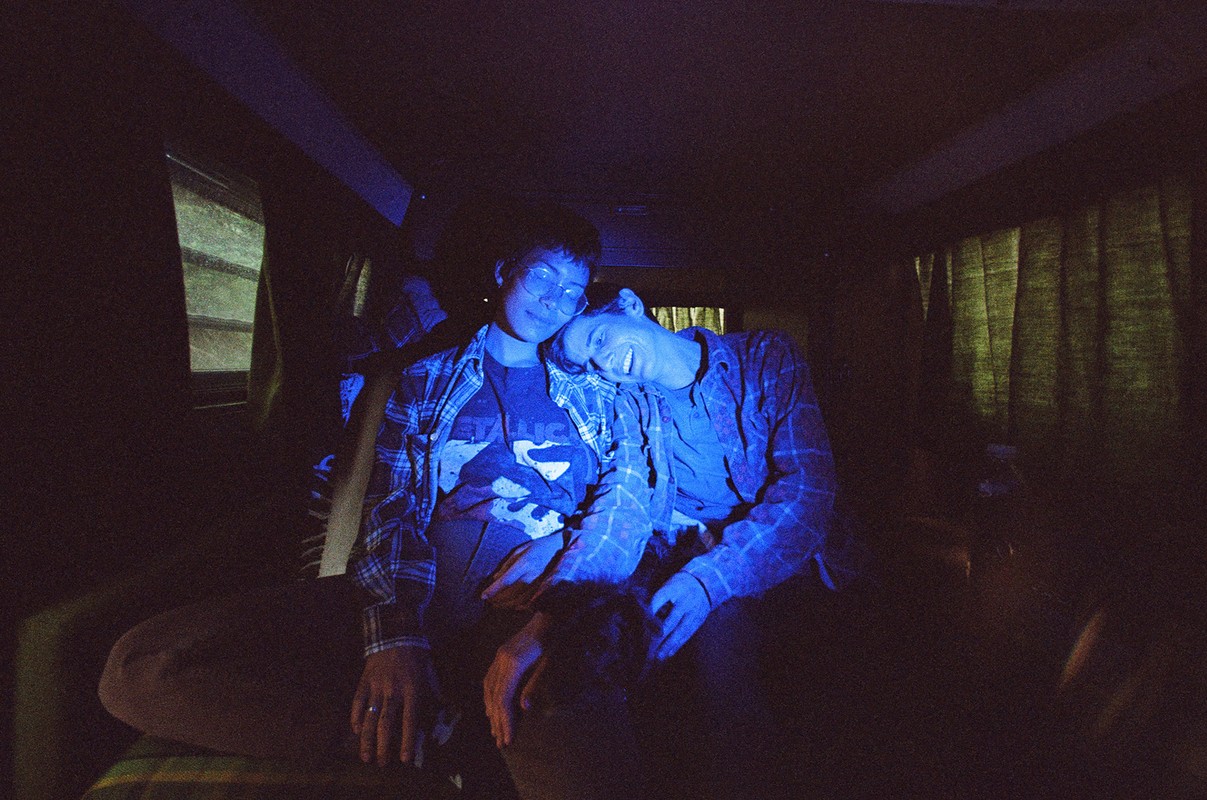 Two people embrace intimately inside a van bathed in blue light, creating a cozy and intimate atmosphere in a dimly lit space.