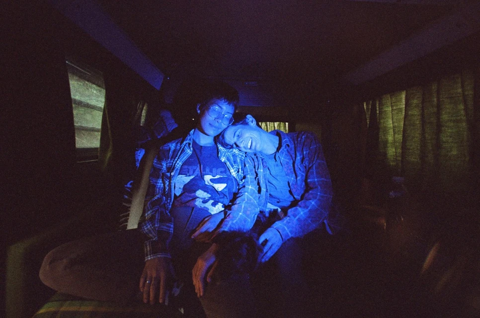 Two people embrace intimately inside a van bathed in blue light, creating a cozy and intimate atmosphere in a dimly lit space.