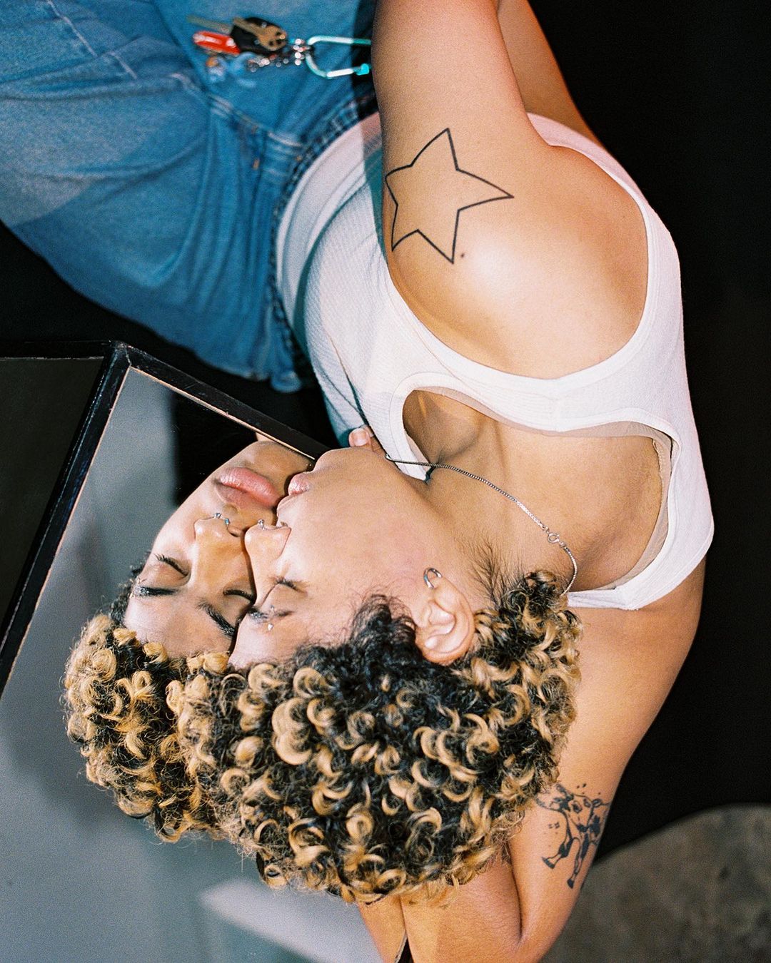 Portrait of a young woman with curly hair gazing into a mirror, wearing a white top and jeans, with a star tattoo on her arm.