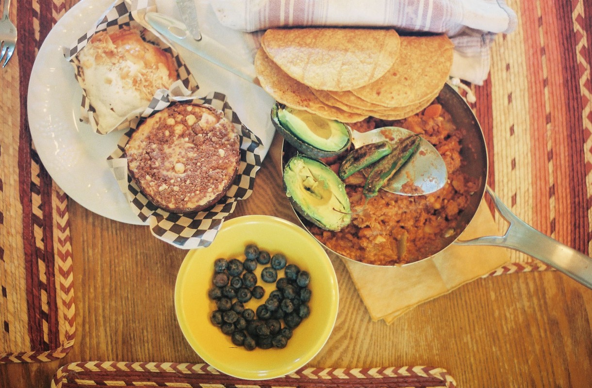 Overhead shot of a pan filled with food, garnished with avocado slices and green chilis, plus tortillas, muffins, and blueberries.