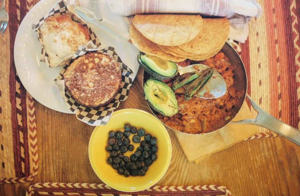 Overhead shot of a pan filled with food, garnished with avocado slices and green chilis, plus tortillas, muffins, and blueberries.