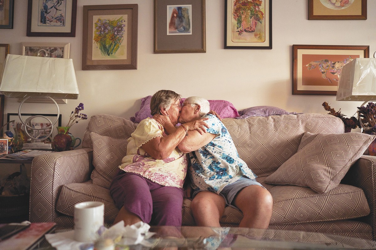 Two elderly adult females share a kiss on a patterned couch inside a home. Paintings hang on the wall behind them. 