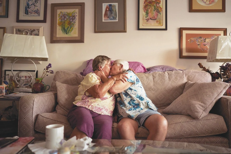 Two elderly adult females share a kiss on a patterned couch inside a home. Paintings hang on the wall behind them. 
