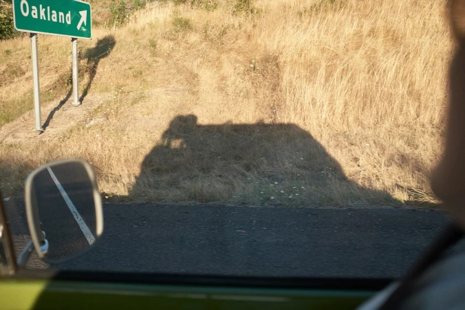 View from a car window of a sign for Oakland on a sunny day, with a dry grassy roadside hill casting a shadow.